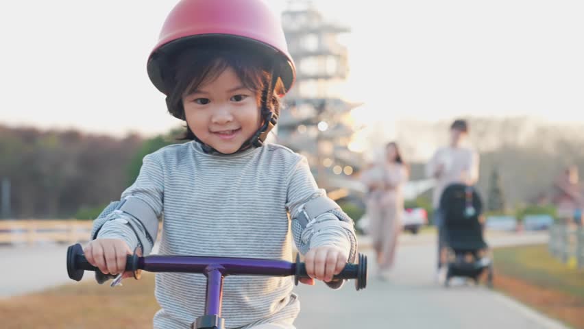 Playful Asian child riding on a bike on the roadside. Girl in helmets cycling on the autumn l. Active smiling children on bikes with parent watching from behind, let kid have their own freedom | Shutterstock HD Video #1100083959