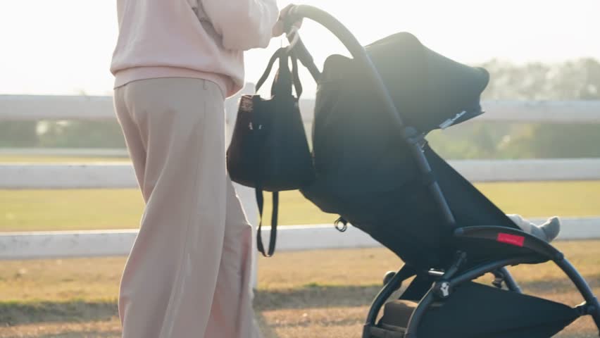 Shot of mother pushing baby stroller car walking around the public area during sunset with beautiful sunlight leak, warm family vibe, new mother amd how to raise a child, modern gear for convenience Royalty-Free Stock Footage #1100085063