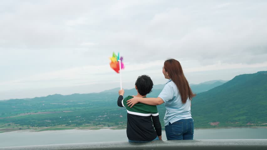 A progressive woman and her son are on vacation, enjoying the natural beauty of a lake at the bottom of a hill while the boy carries a toy windmill.