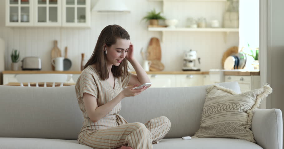 Pretty girl puts in wireless earphones using smart phone, relaxing on sofa. Young generation and modern technologies usage for leisure, easy and comfortable devices usage, pastime at home on internet | Shutterstock HD Video #1100087689