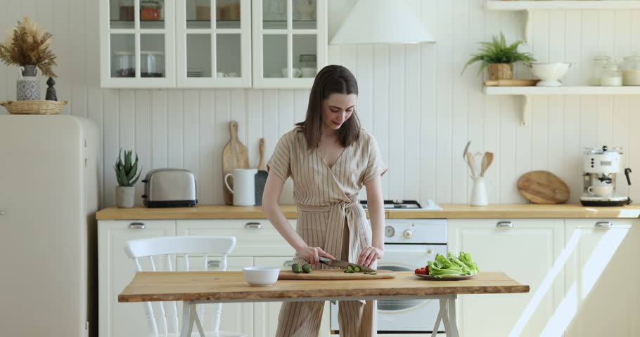 Pretty slim woman holds knife, cuts fresh cucumber, prepare vegetarian salad, keep diet, making natural dish standing alone in cozy domestic kitchen. Cooking process, lifestyle, chores, healthy eating | Shutterstock HD Video #1100087725