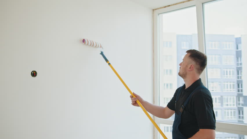 Painter man. A bearded construction worker in a blue overalls is painting a wall in the hallway. He uses a roller. Man painting wall in her new apartment. Renovation and redecoration concept Royalty-Free Stock Footage #1100092113