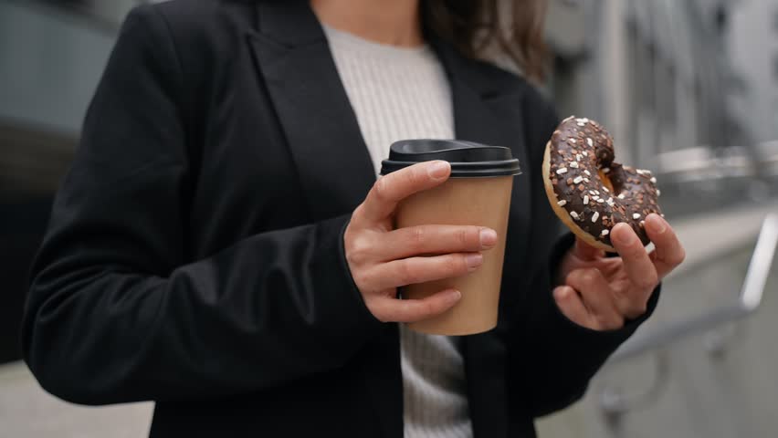 Young Caucasian with brown hair wearing black jacket drinking coffee and eating chocolate donut between skyscrapers . Outdoor, snacks time.