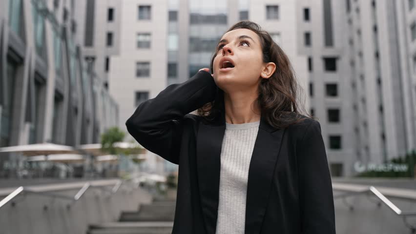 Young Caucasian female with dark long hair walking in city center around grey skyscrapers, girl likes view of high contemporary architecture.
