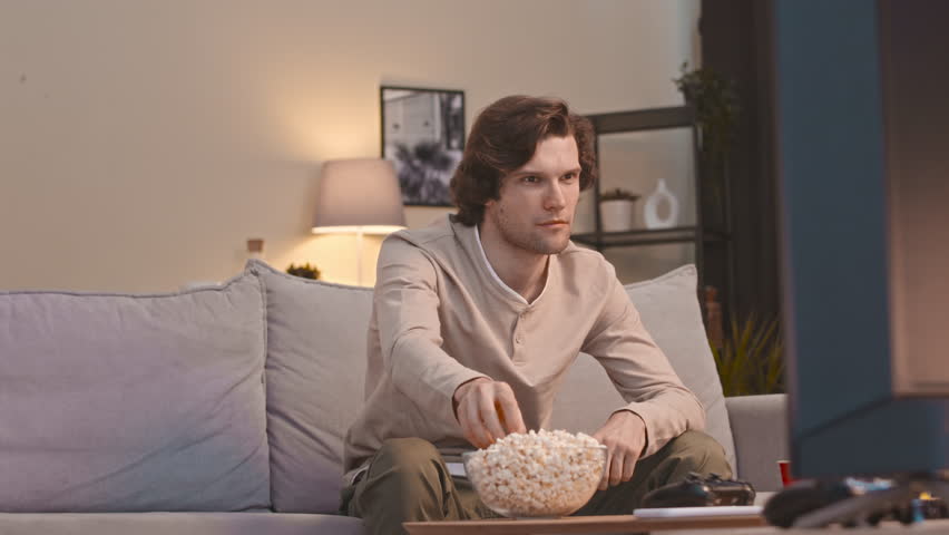 Medium shot of young Caucasian man eating popcorn and laughing while watching hilarious comedy film on TV sitting on couch in living room in evening