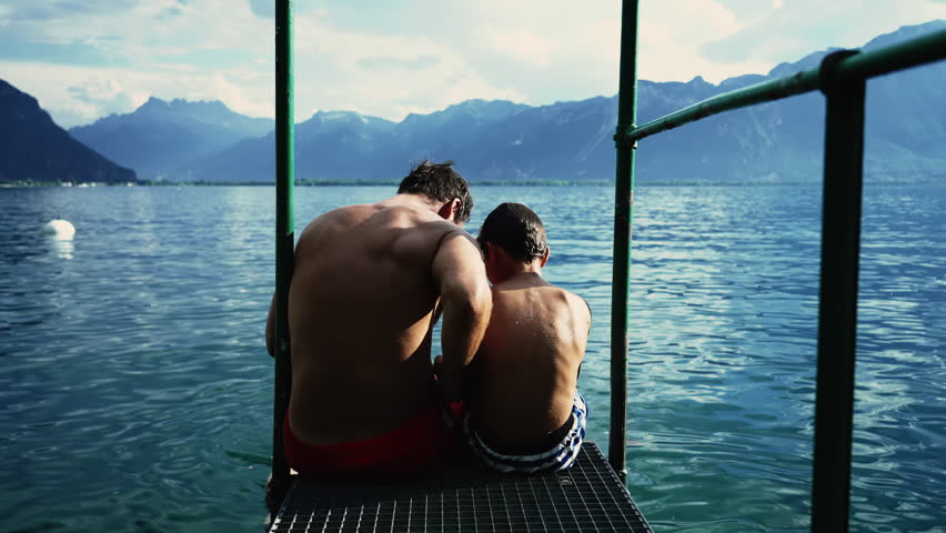 Father and son sitting by pier overlooking scenic beautiful lake view with mountains in background. Dad and son bonding together by lake Geneva