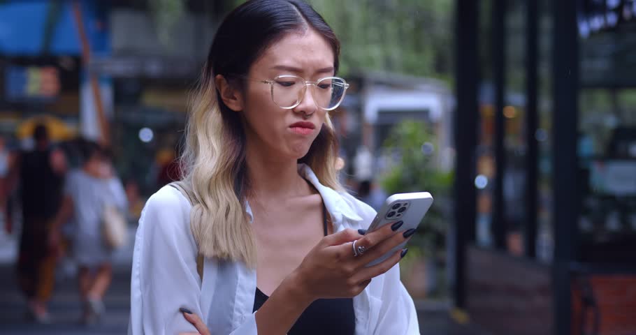 Portrait of sad displeased young asian woman looking at mobile phone screen, dissatisfied with bad news message, spam or scam sms. Unhappy sad asian female using mobile phone feel stress in city Royalty-Free Stock Footage #1100123059