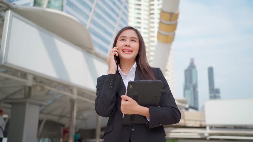 An attractive Asian businesswoman talking on a smartphone with a smile walks in outdoor urban city buildings. Asian lady chats with her friend using a mobile phone to listen to the business news. | Shutterstock HD Video #1100127401