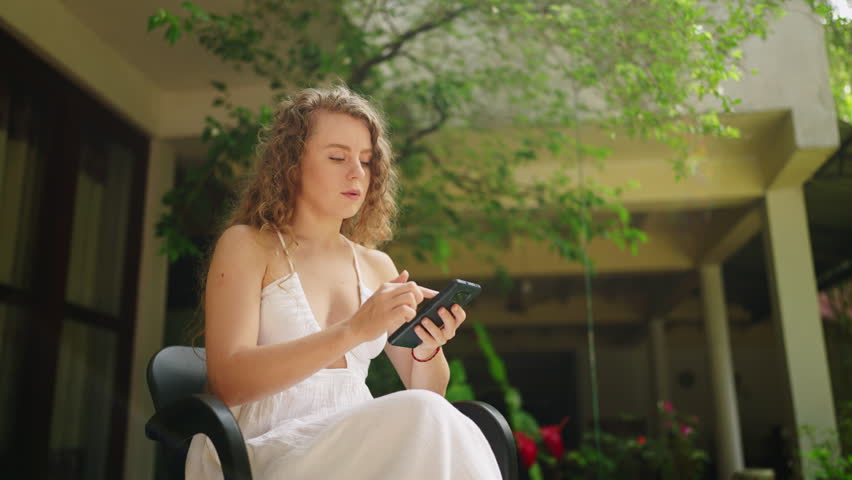 Young woman using smart phone texting with AI, surfing social media, checking news, playing mobile games or texting messages sitting under tree. Millennial lady spending time with cell gadget outside