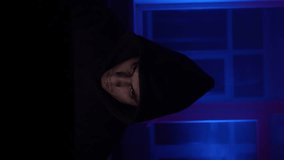 Vertical Video, Hooded Male Hacker Sits Working at a Laptop Cracking a Security Code at Night in Neon Lighting. Concept of Cyberspace and Cybercrime. Slow Motion.
