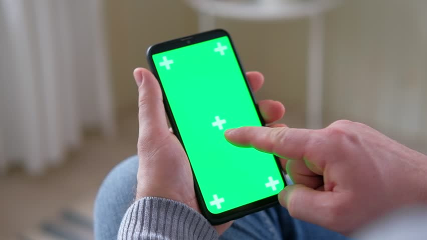 Young man sitting at home holding smartphone green mock-up screen in hand. Male person using chroma key mobile phone. Vertical mode. Touching, swiping display, tapping, surfing Internet social media | Shutterstock HD Video #1100132791