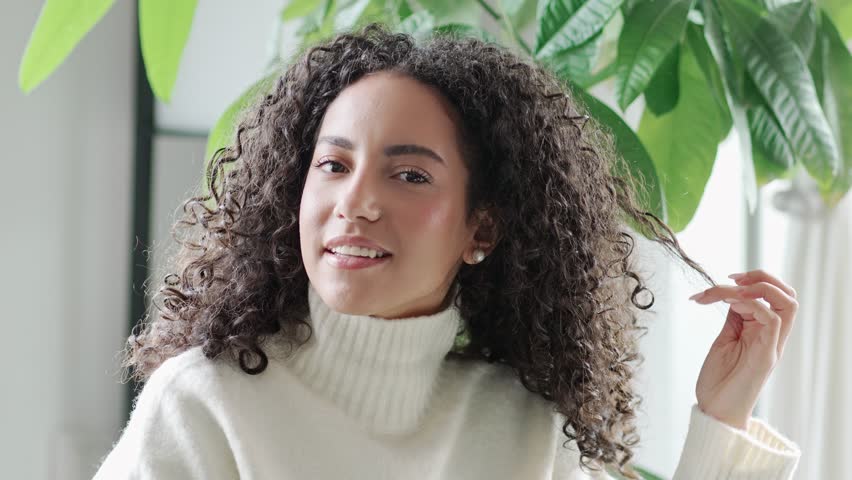 Young pretty hispanic woman with white teeth dental smile relaxing at home. Happy stylish relaxed latin beautiful positive lady playing with curly hair looking at camera. Close up portrait. | Shutterstock HD Video #1100134585