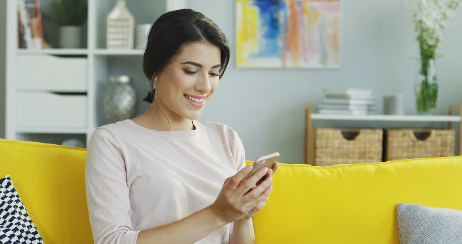 Beautiful and joyful young woman holding a smartphone, reading or watxhing something and laughing. At home. | Shutterstock HD Video #1100135497