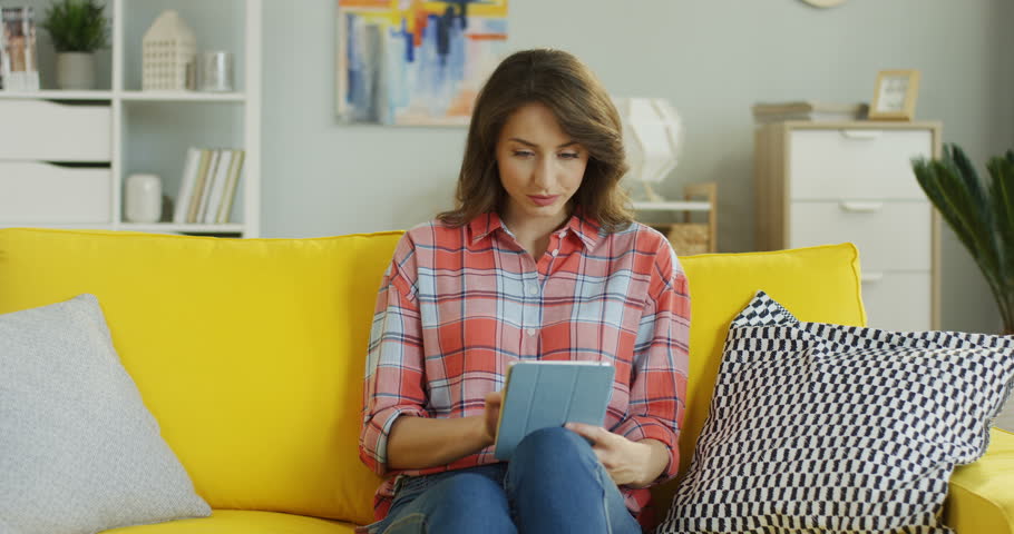 Young attractive woman being surprised from something she seeing on the tablet device screen while sitting on the yellow couch at home. | Shutterstock HD Video #1100135613