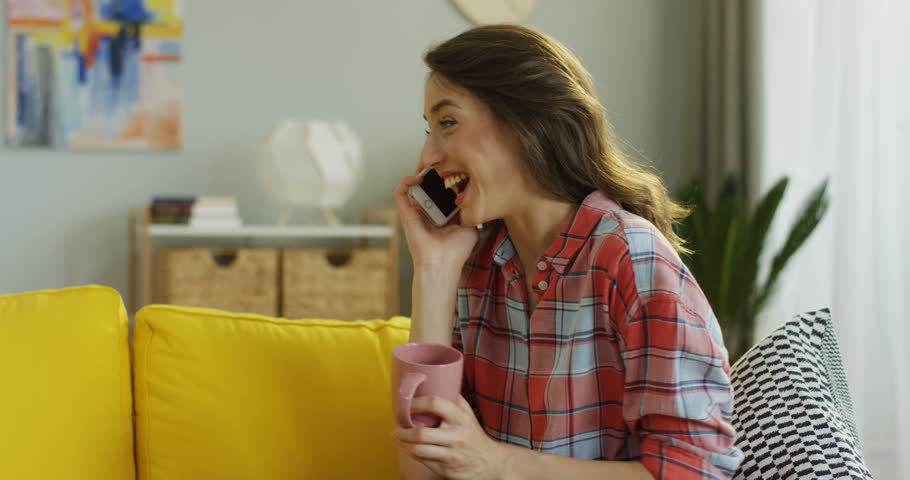Young pretty woman sitting in the living room at home and talking cheerfully on the phone while holding a cup in a hand. | Shutterstock HD Video #1100135621