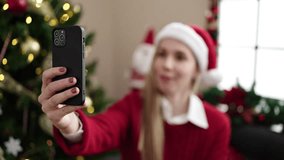 Young blonde woman having video call sitting on sofa by christmas tree at home