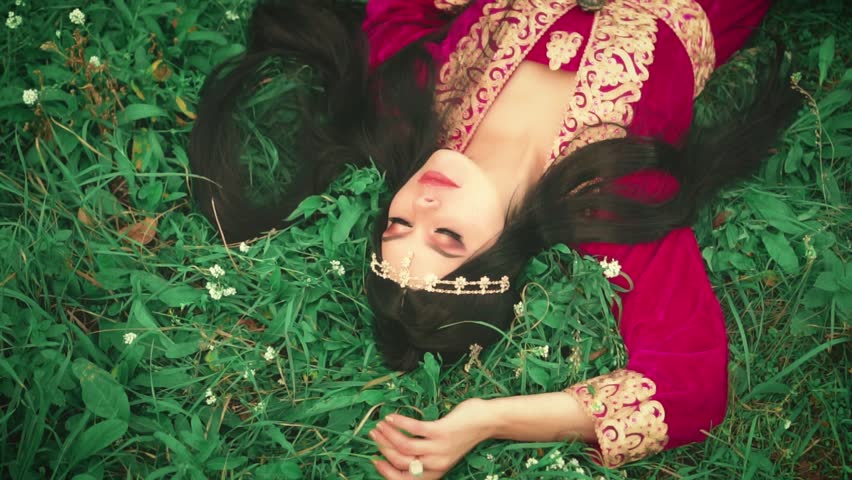 Happy fantasy woman lies on green spring grass, sleeps sees sweet dream. Spring nature white flowers glade. fairy tale sleeping beauty princess girl, long dark hair, crown on head. oriental red dress