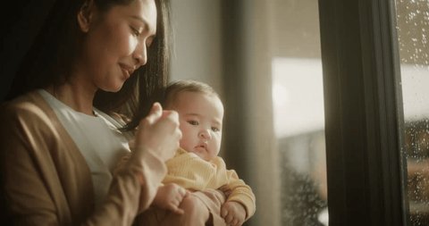 Beautiful Young Asian Woman Holding her Baby in her Arms While Standing Next to a Window at Home. Cute Little Toddler Resting in Her Mother's Embrace as She Affectionately Caresses her Hair: stockvideo