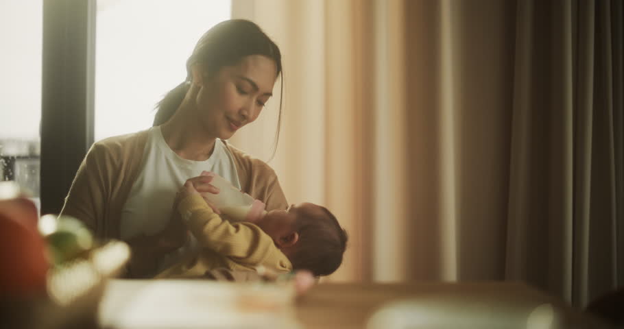 Portrait of Beautiful Asian Mother Feeding Milk to her Infant Using a Baby Bottle at Home. Young Woman New to Motherhood Bonding with her Infant and Enjoying an Affectionate Moment Royalty-Free Stock Footage #1100144523