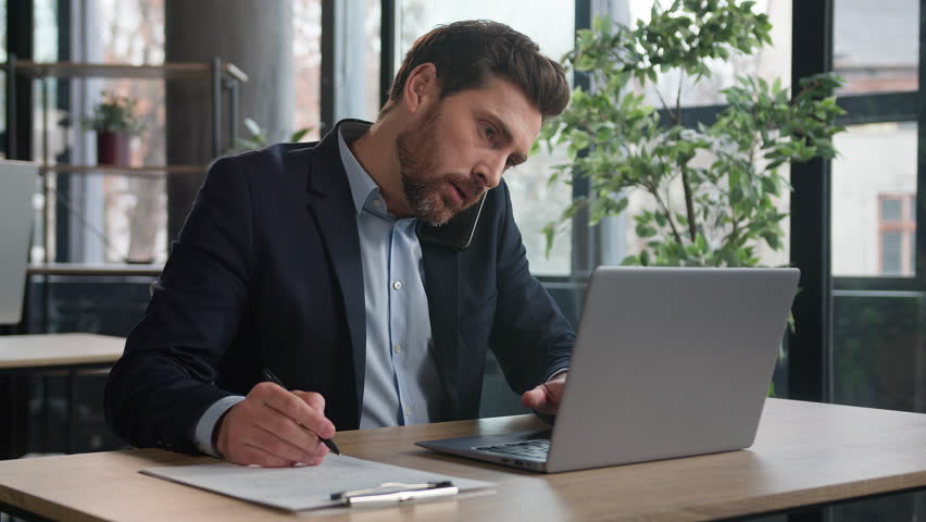 Busy successful caucasian adult businessman in office typing on laptop talking to client on mobile phone writing notes explain condition multitasking man manager communicate on cellphone in workplace | Shutterstock HD Video #1100145175