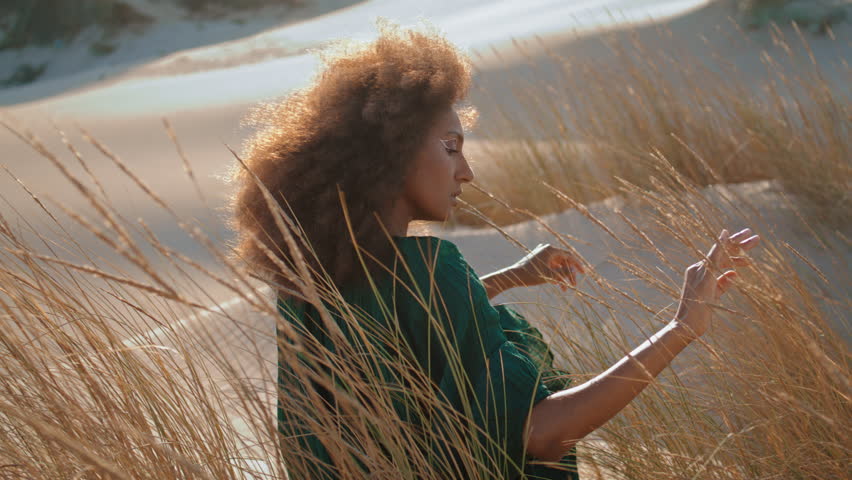 Dreamy young woman enjoy desert nature sitting on sand touching dry grass swaying wind close up. African american girl wearing black dress relaxing under soft summer sunlight near dunes wilderness.