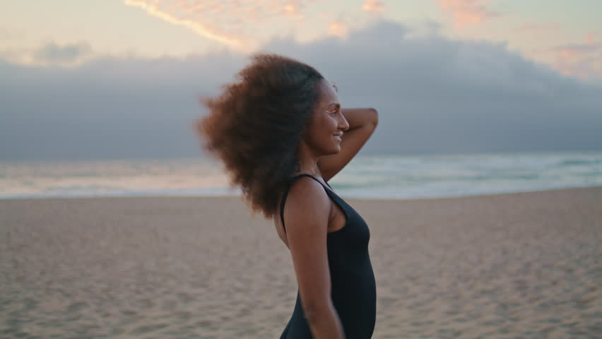 Relaxed woman tourist walking sandy gloomy beach in black swimsuit at summer dusk. Carefree african american girl relaxing on beautiful seashore at twilight. Attractive young brunette going near sea. Royalty-Free Stock Footage #1100146547