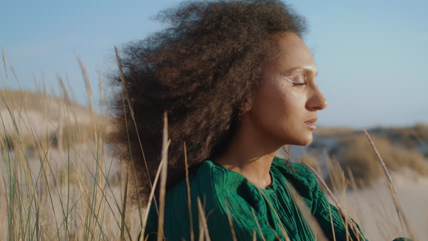 Portrait of african american young woman sitting alone on summer desert looking camera. Dreamy attractive girl wearing black dress enjoying beautiful landscape. Wind blowing on curly dark hair closeup
