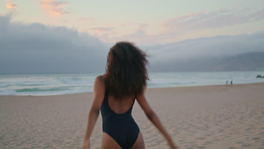 Excited african american girl running on seashore raising hands up at overcast evening. Curly happy woman in black swimsuit jumping spinning have fun at empty beach. Attractive brunette smiling. Royalty-Free Stock Footage #1100146559