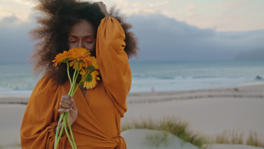 Portrait of happy african american girl posing on sand seashore with orange flowers. Attractive cheerful woman sniffing cute bouquet standing on beach at overcast windy evening. Curly brunette smiling