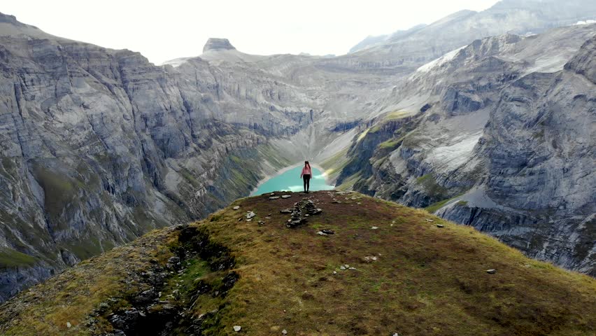 A flyover above a hiker enjoying the view over lake Limmernsee in Glarus, Switzerland, the turquoise colored water of which is surrounded by tall Swiss Alps peaks and cliffs