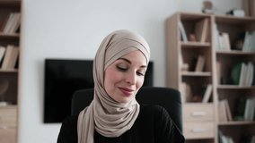 Smiling positive confident muslim woman in formal wear and hijab sitting at office chair. Company worker with headscarf looking at camera indoors. Concept of people and business.