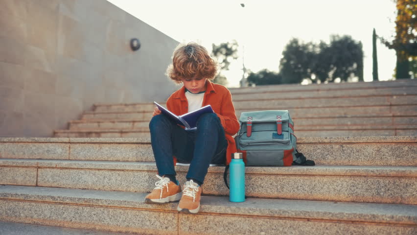 Back to school. A cute child with a backpack sits on the stairs near the school and reads a textbook. Elementary school student studying homework after class. Royalty-Free Stock Footage #1100157585