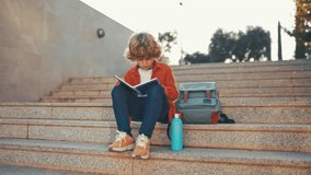 Back to school. A cute child with a backpack sits on the stairs near the school and reads a textbook. Elementary school student studying homework after class. - Powered by Shutterstock - Get 15% off with code: PIKWIZARD15