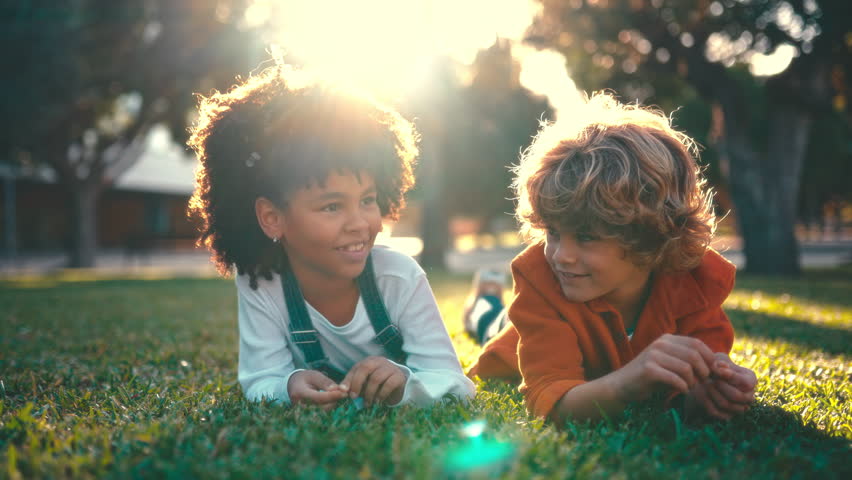 Adorable children lying on the grass in the park. African American girl with afro hair and caucasian stylish boy relaxing and playing, having fun on a day off. School break.