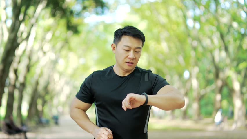 Asian runner jogging in an urban city park and looking smart watch. Man using smartwatch bracelet. Portrait of fitness man checking result. A handsome male in a sports suit enjoys running outdoors