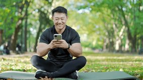 Happy asian man in tracksuit sitting on mat and browsing smartphone in urban city park. A handsome male is smiling while reading messages from participants in an online yoga class that was just held - Powered by Shutterstock - Get 15% off with code: PIKWIZARD15