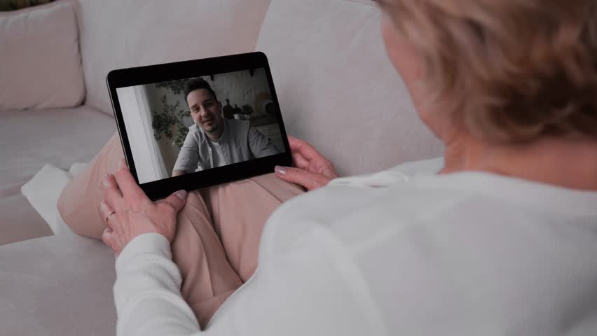 An adult woman communicates with her son on a video call using a tablet while sitting in a cozy warm apartment on the couch. Living away from your parents. High quality 4k footage Royalty-Free Stock Footage #1100160421