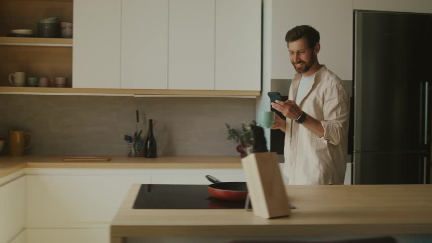 Happy Caucasian man going to window in modern interior apartment sunny kitchen enjoy morning coffee scrolling news feed on phone. Business man using smartphone
in his home office.  Royalty-Free Stock Footage #1100160953