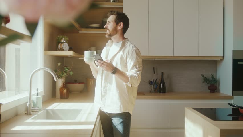 Happy Caucasian man going to window in modern interior apartment sunny kitchen enjoy morning coffee scrolling news feed on phone. Business man using smartphone
in his home office.  | Shutterstock HD Video #1100160953