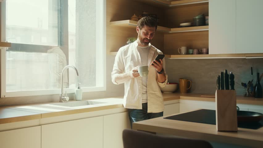 Caucasian man standing near window in modern interior apartment sunny kitchen having morning coffee scrolling news feed on phone. Business man using smartphone
in his home office.  Royalty-Free Stock Footage #1100160961