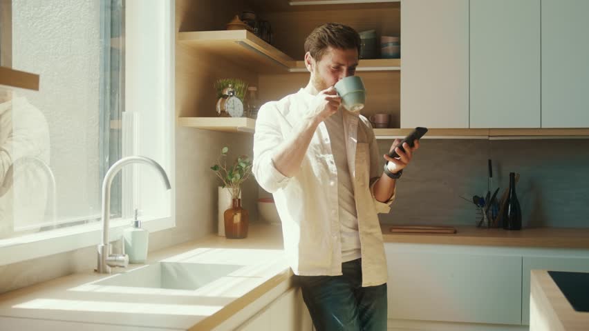 Caucasian man standing near window in modern interior apartment sunny kitchen having morning coffee scrolling news feed on phone. Business man using smartphone
in his home office. 