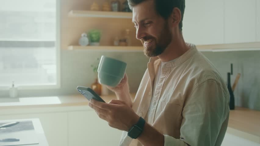 Happy Caucasian man sitting on chair in modern interior apartment sunny kitchen enjoy morning coffee scrolling news feed on phone. Business man using smartphone
in his home office.  Royalty-Free Stock Footage #1100160969