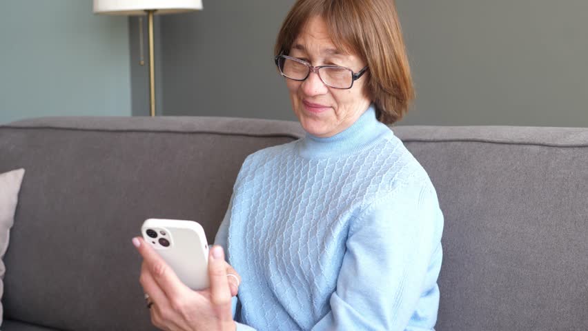 Smiling adult lady uses a smartphone to type while sitting at home on a sofa in a cozy house. Elderly people and modern technologies. Baby Boomers Royalty-Free Stock Footage #1100161691