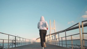 Back view young slim woman in sport hoodie and black leggings jogging by running track on city bridge in morning sun light enjoy active hobby outdoors. Follow shot. Slow motion Handheld - Powered by Shutterstock - Get 15% off with code: PIKWIZARD15