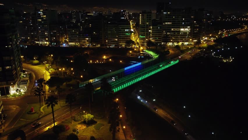 Night time 4k footage of a bridge being lit with LED color changing lights. Many city lights shine in distance. Many tall buildings in the background. Located in Miraflores district of Lima, Peru