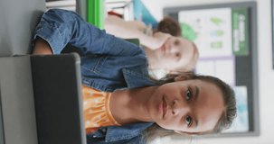 Vertical video of happy bircacial schoolgirl sitting at desk, using tablet in class, copy space. Education, childhood, inclusivity, technology, communication, school and learning concept.