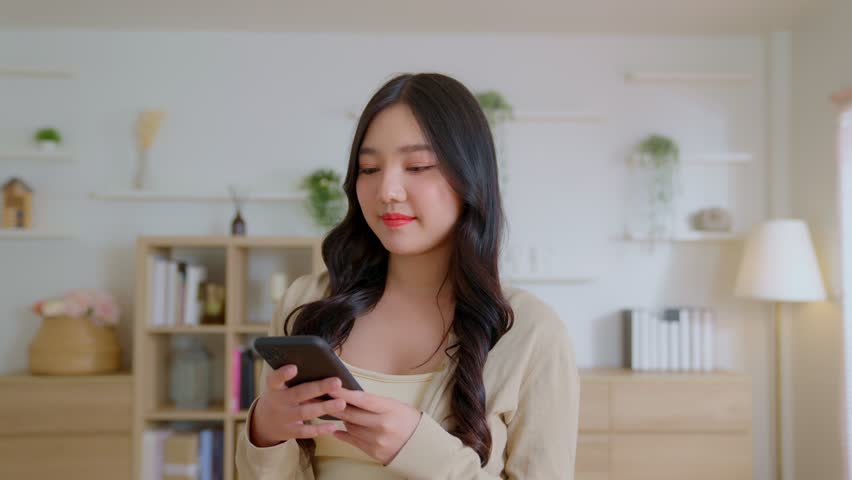 Attractive beautiful Asian woman happy playing with her mobile phone in the living room of new home. Female looks at the camera and smiles happily and very cute. Asia girl close up front portrait | Shutterstock HD Video #1100171681