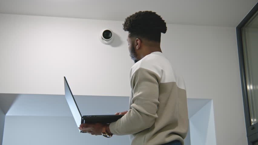 African american man sets up security cameras in office corridor using laptop. Installer checks CCTV cameras in computer program. Monitoring system. Concept of surveillance system and privacy. Zoom in Royalty-Free Stock Footage #1100174103