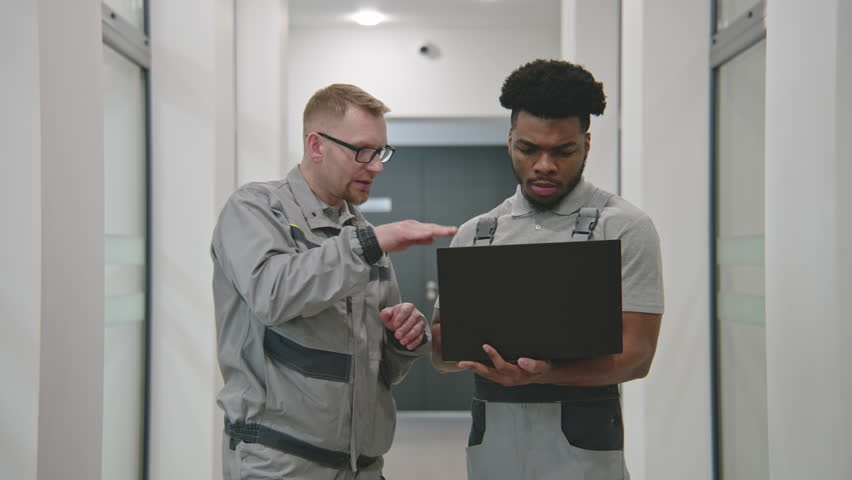 Two installers in uniform stand in corridor and discuss security cameras installation in business office. Men set up CCTV cameras using software on laptop. Concept of surveillance system. Dolly zoom. Royalty-Free Stock Footage #1100174127