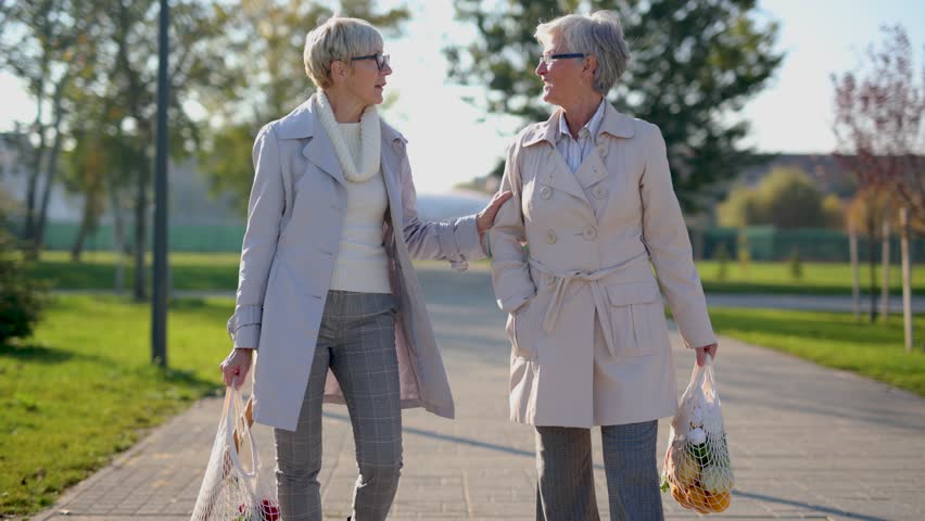 Two senior female friends are holding grocery bags and walking through park | Shutterstock HD Video #1100177289